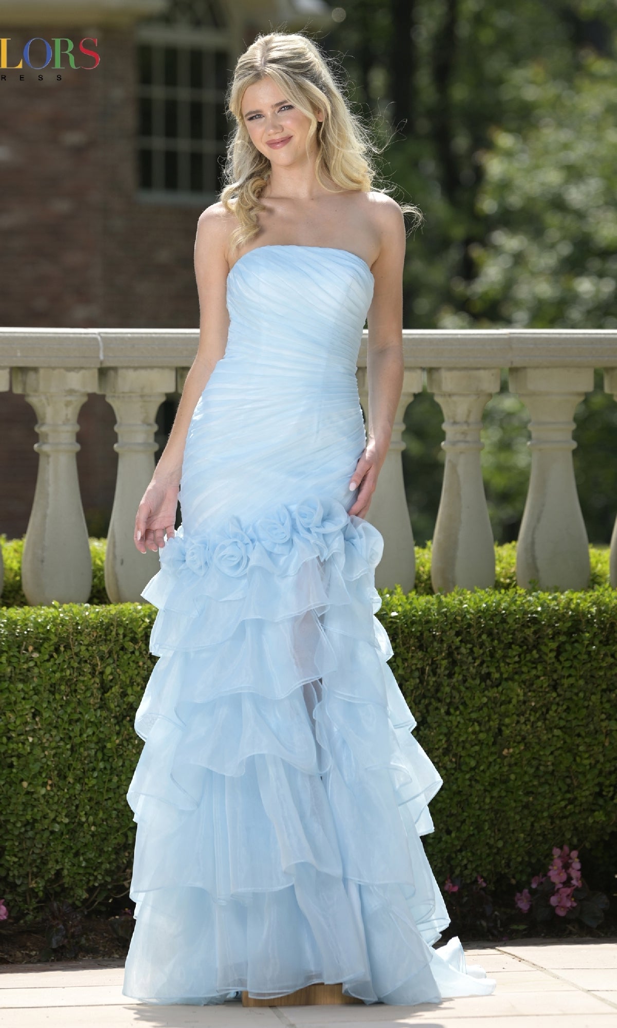 A woman in the Colors Dress 3882 long prom dress, featuring a strapless ruched fitted bodice and light blue ruffled gown, stands outdoors by a stone railing and greenery.